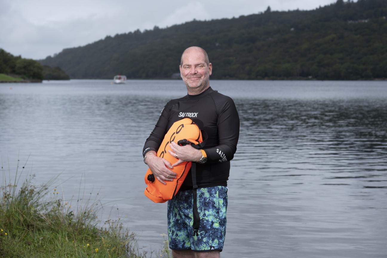 A man standing on the shore of lake wearing a wetsuit and holding a float.