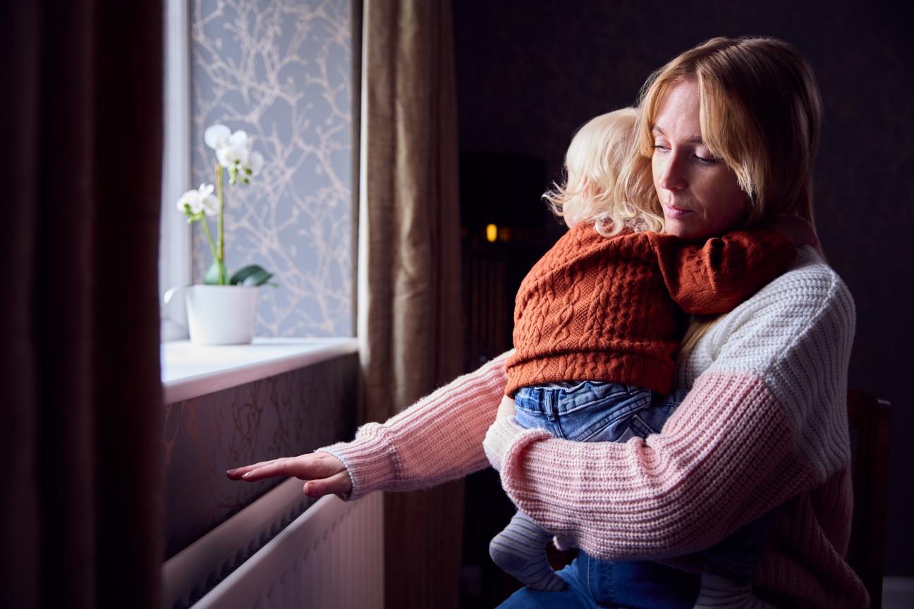 A woman holding a baby feels for heat from a radiator
