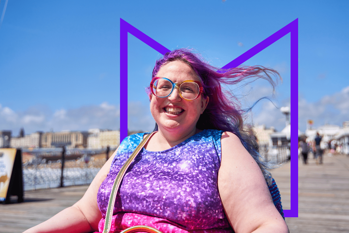 A woman in a wheelchair on a pier on a sunny day. She's smiling at the camera and her purple-dyed hair is blowing in the wind.